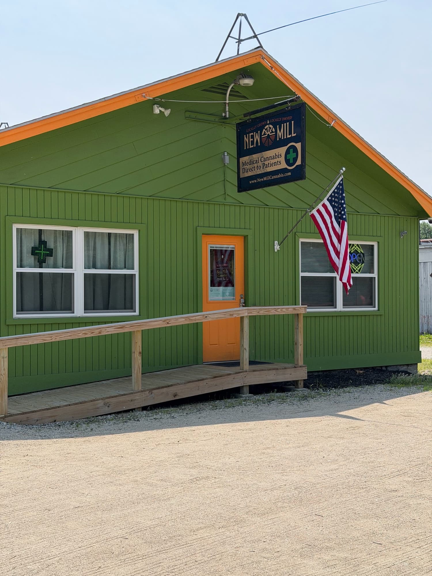 The front of the green New Mill shop with its sign, an American flag and a wooden ramp up to the orange door.